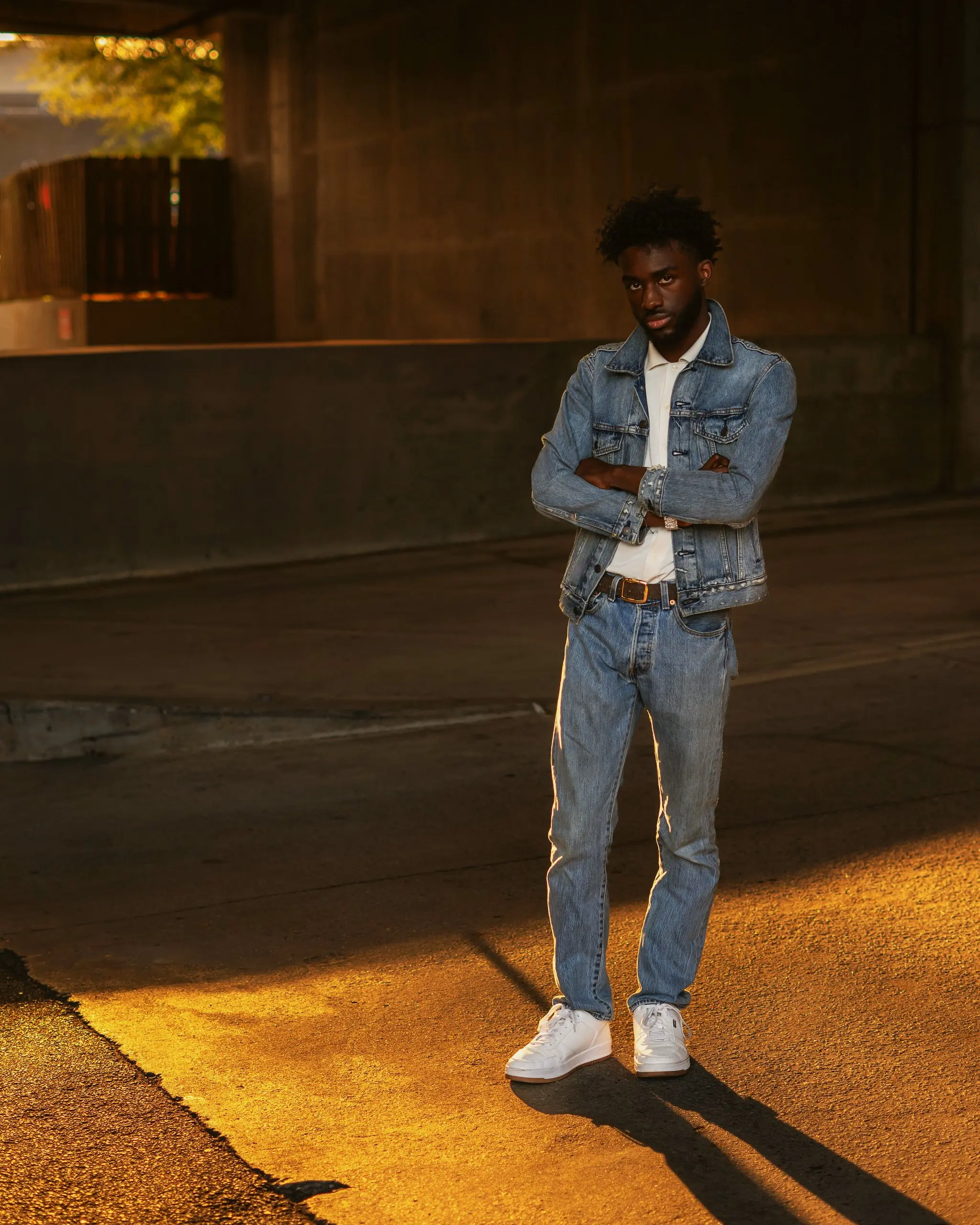 Washed denim jacket and jeans on a young Black man standing in low sun under an overpass, long shadow