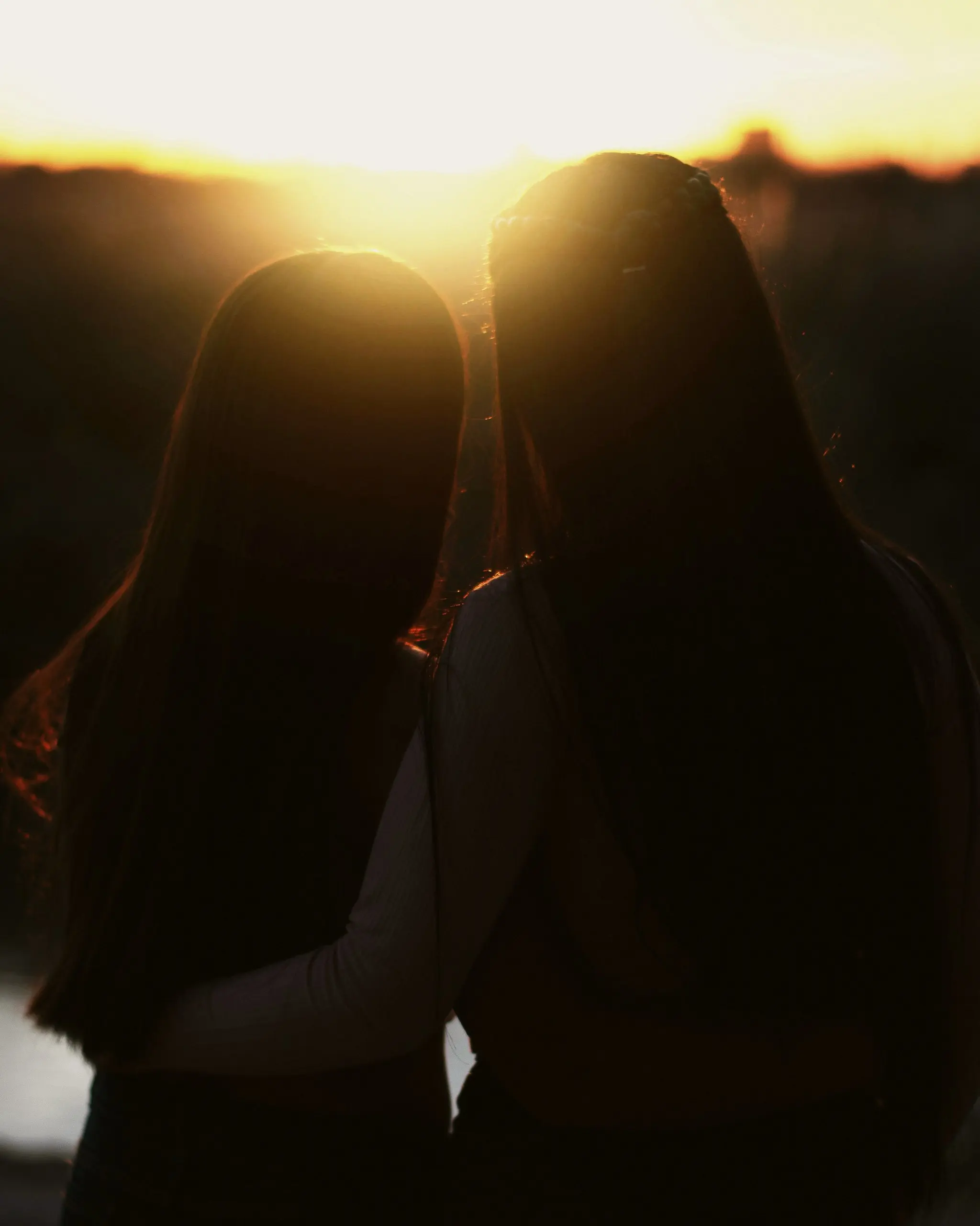 Dark tops on two friends seen from behind, arm in arm, silhouetted against a golden sunset