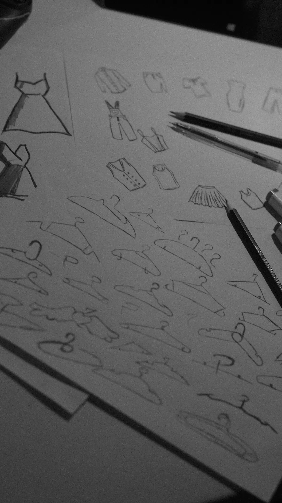 Pencil sketches of garments and hangers on paper, with pencils on the page